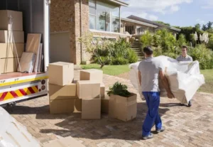 Movers loading boxes into a truck while homeowners and a dog stand nearby