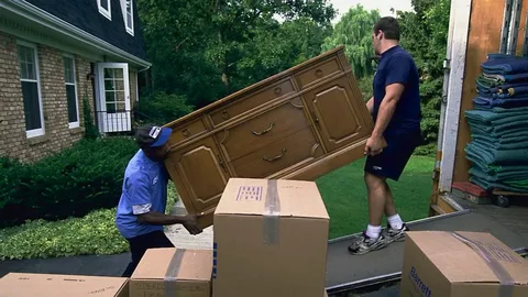 Two movers carrying a wooden dresser into a moving truck.